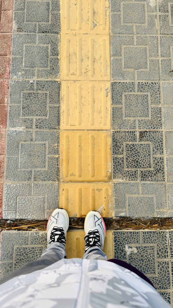 A top-down view of a person in white sneakers standing on a sidewalk with yellow tactile tiles, surrounded by gray patterned pavement.