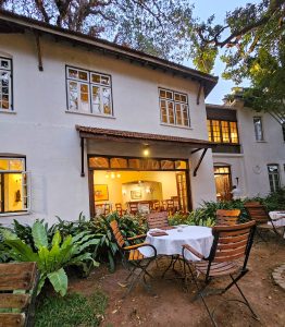 An elegant white colonial-style building featuring an outdoor patio with wooden chairs and white-clothed tables, illuminated by warm indoor lighting.
