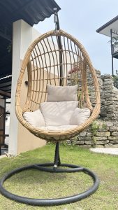 Outdoor hanging rattan egg chair with beige cushions on a metal stand, placed on a grassy lawn beside a stone wall and modern house exterior.