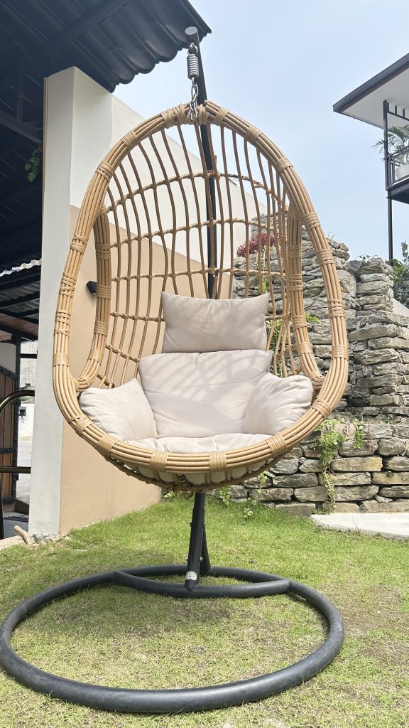 Outdoor hanging rattan egg chair with beige cushions on a metal stand, placed on a grassy lawn beside a stone wall and modern house exterior.