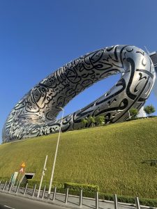 Silver, hollow, ring-shaped building covered in black Arabic calligraphy, rising from green grass against a blue sky.
