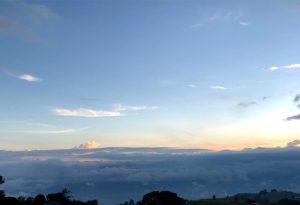 A panoramic view of a cloudy sky during twilight, featuring layers of dark clouds and hints of pink and orange hues as the sun sets. The upper part of the sky is a clear blue, with wispy clouds scattered throughout. In the foreground, silhouettes of trees and hills are visible, enhancing the serene landscape.