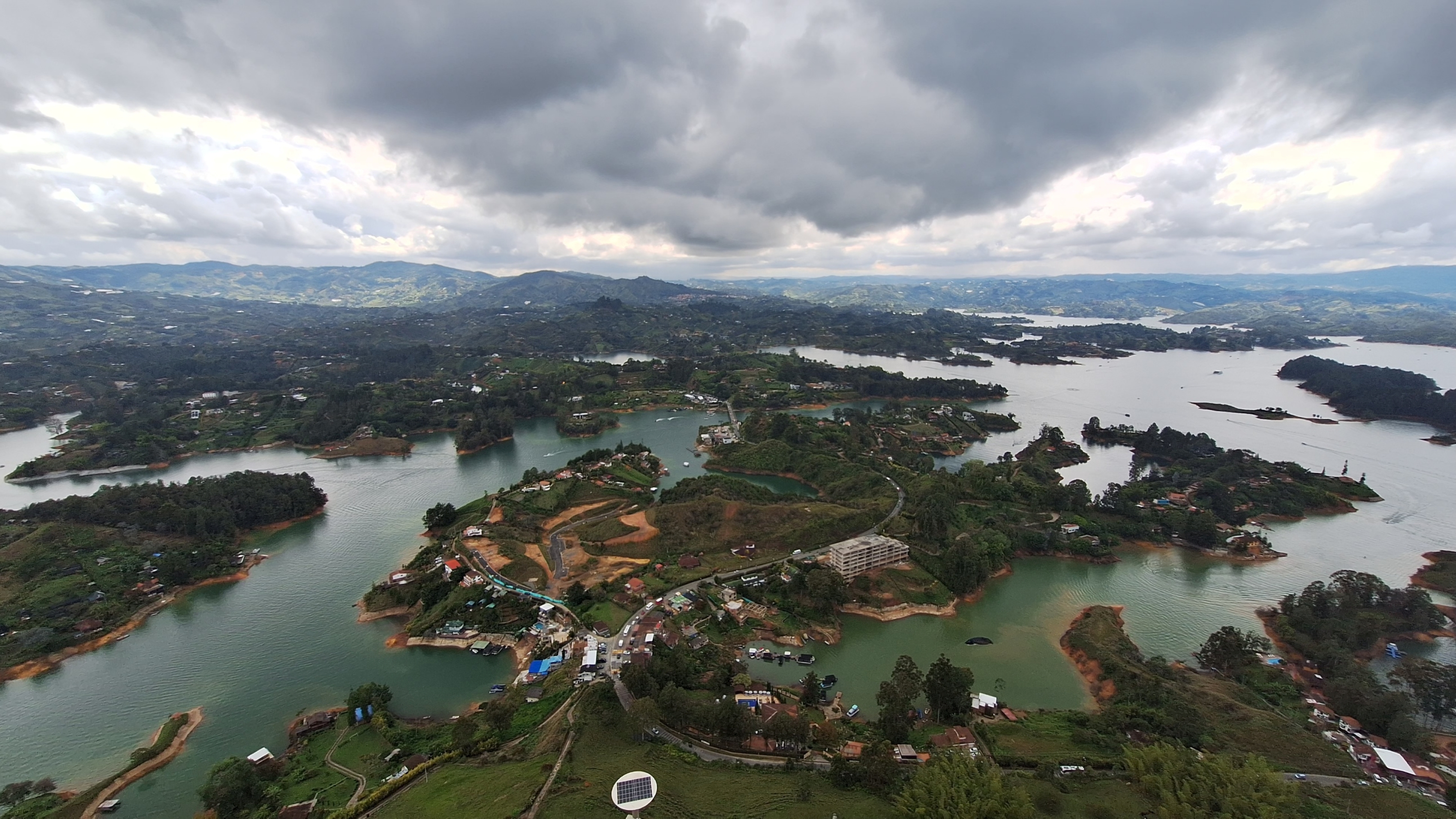 An aerial view of a lush, green landscape featuring multiple lakes and islands. The scene is set against a backdrop of rolling hills and a cloudy sky, creating a serene and picturesque environment. Various small villages with houses and roads are scattered throughout the landscape, and the water reflects the overcast sky. Piedra del Peñol, Guatapé.