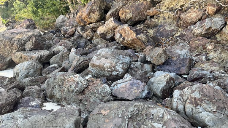 A close up view shows a pile of large, rough rocks on a beach. The rocks have different colors including brown, grey, and reddish tan. Some rocks have sharp edges and deep cracks. In the background, a few green trees are visible at the edge of the pile.