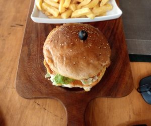 A chicken burger with lettuce on a sesame bun topped with a black olive, served on a wooden board with chips in the background.