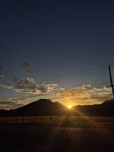 A golden sunset shining behind mountain silhouettes under a clear blue sky, with a quiet road and fence in the foreground