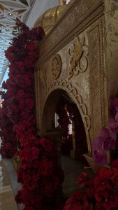 An ornate golden structure with intricate designs, partially framed by vibrant red flowers, with soft lighting highlighting its detailed textures and decorative patterns.