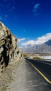 A winding road bordered by a rocky cliff on the left side, with a blue sky and scattered clouds above. In the distance, majestic mountain ranges are visible, some peaks capped with snow.
