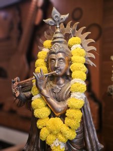 A detailed Krishna statue playing flute, decorated with a yellow flower garland. A warm, devotional portrait captured at Mumbai Airport.