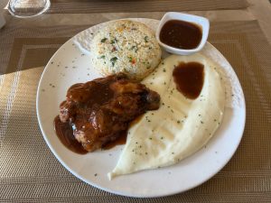 A plate of food featuring grilled chicken in sauce, creamy mashed potatoes, and colorful fried rice. A small dish of extra sauce is on the side.