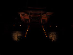 A dimly lit pathway, lined with soft, glowing candles, leads toward a traditional building. The warm light creates an inviting, mystical atmosphere that highlights the structure's ornate architectural details and creates dramatic shadows against the stone wall and garden.