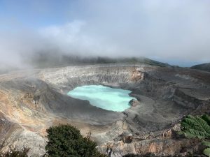 This image shows a volcanic crater on a cloudy day, with a light blue lake in the center surrounded by rocky terrain. Mist and low clouds cover part of the area, creating a cool and mysterious atmosphere, while some vegetation can be seen around the edges of the crater.