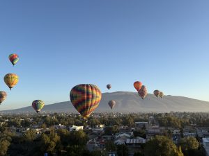 Colorful hot air balloons float in a clear blue sky at sunrise. Each balloon has bright stripes like red, yellow, green, and blue. In the background, there are soft mountains, green land, trees, and small buildings, making the scene calm and beautiful.