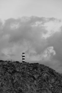 A solitary striped lighthouse stands on a rocky hill under dramatic clouds in a black-and-white, cinematic landscape.