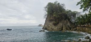 A calm sea under a cloudy sky with a large rock covered in vegetation in the foreground, taken in Quepos.