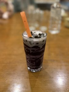 A tall glass of dark bubble tea with ice and tapioca pearls, topped with foam. An orange straw sits in the drink, placed on a wooden table.