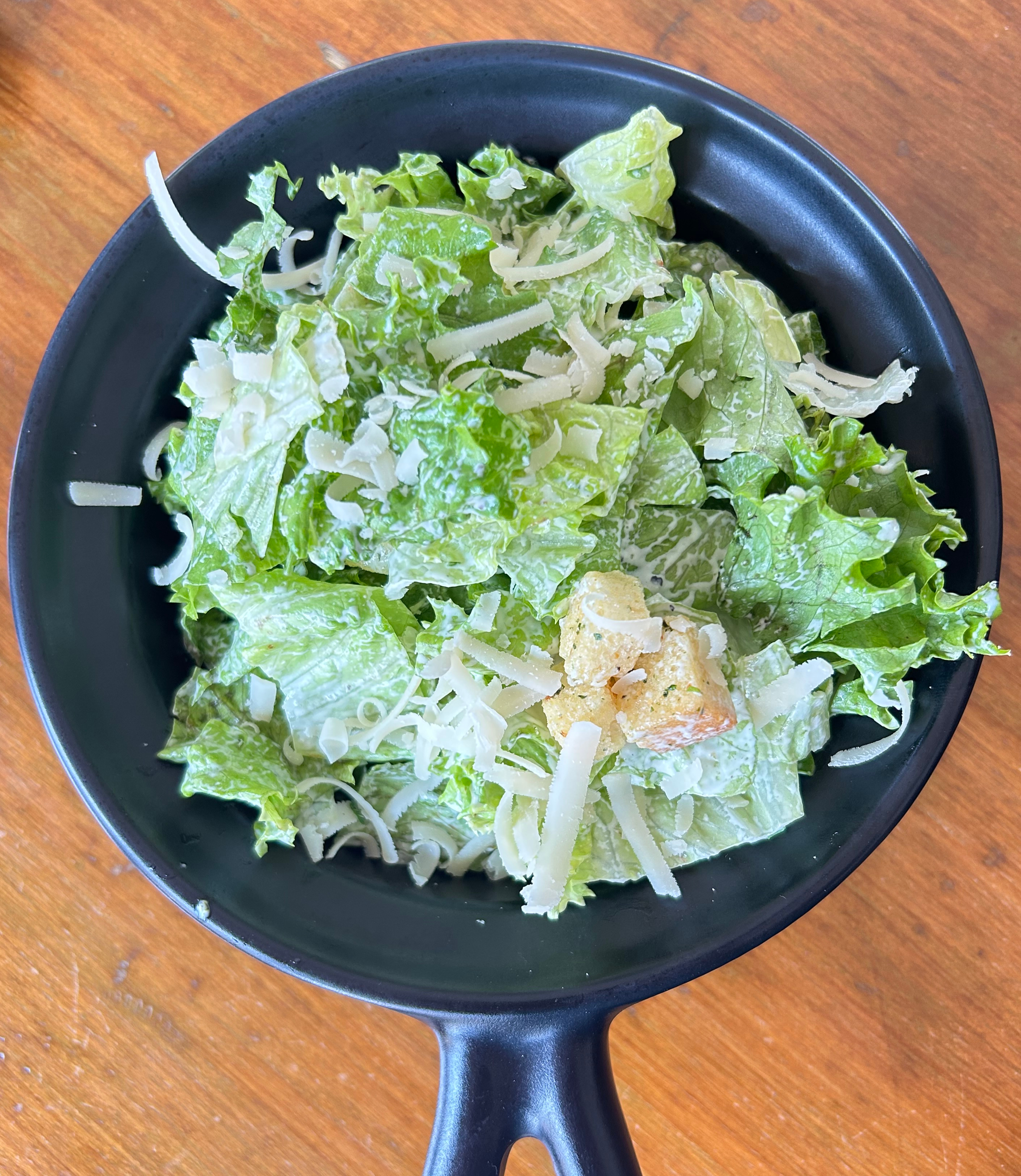 A black bowl sits on a wooden table, filled with a fresh salad.