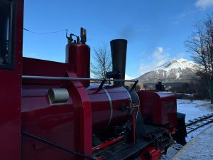A vintage red steam locomotive is positioned in the foreground, featuring a tall black smokestack and a shiny metallic exhaust pipe. In the background, a snow-capped mountain rises against a bright blue sky, with a few wispy clouds.