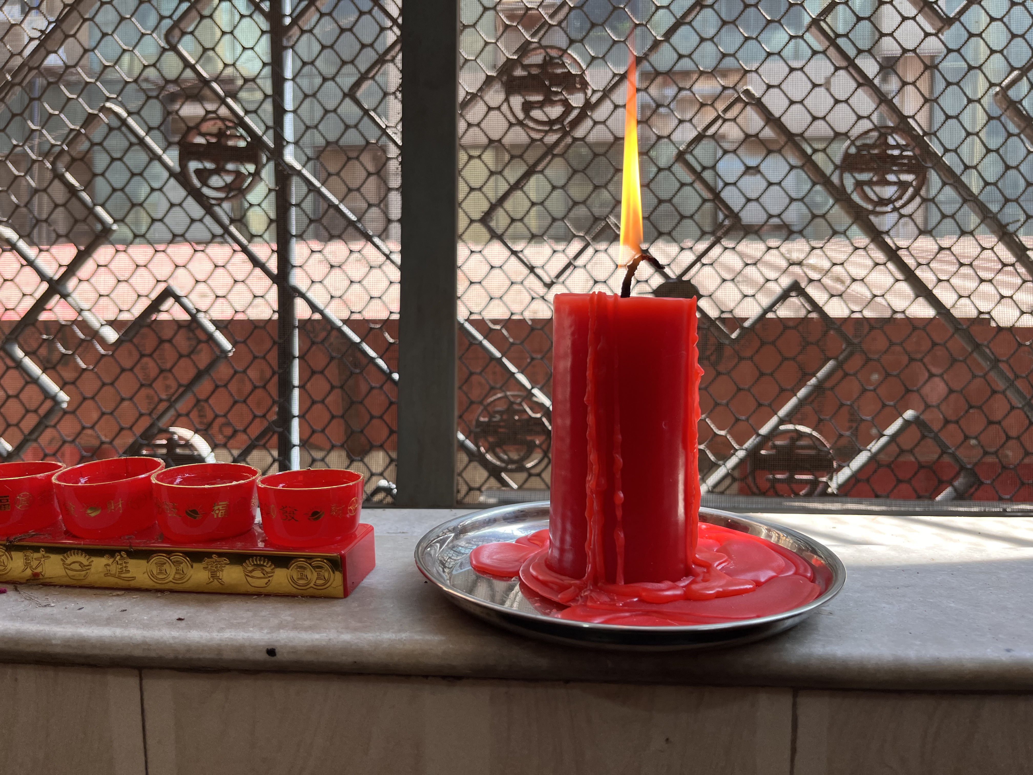 A red candle is lit and melting on a silver plate, with bright wax dripping down. In the foreground, there are four small red cups arranged on a red and gold tray.