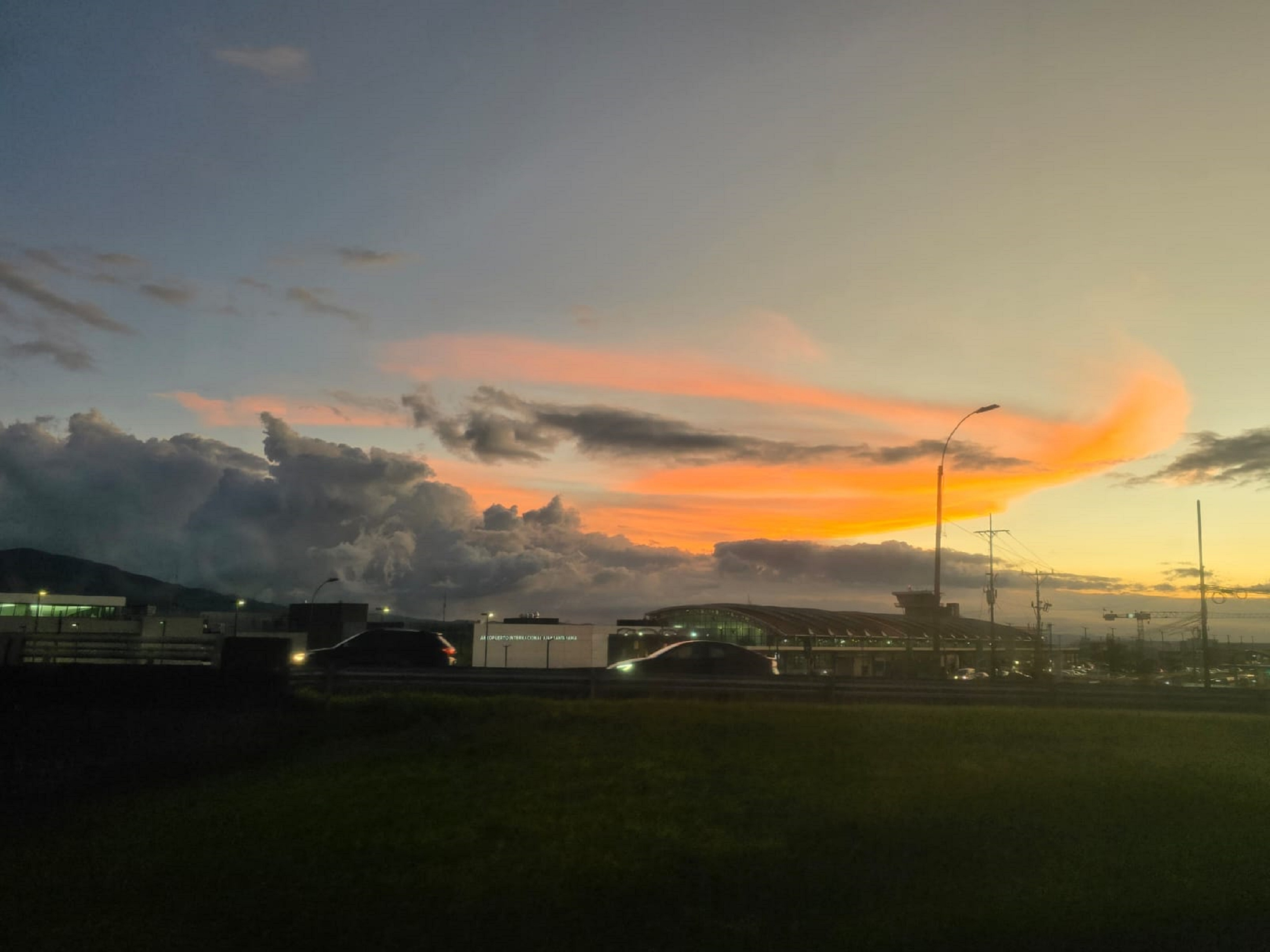 A scenic view of the sky at dusk, showcasing swirling clouds illuminated by shades of orange and pink as the sun sets. In the foreground, there are buildings and a road with lights, while the horizon features distant mountains and various utility poles. The atmosphere conveys a tranquil yet vibrant end to the day.