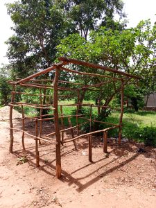 A small wooden shelter stands on red sandy ground, built from rough poles and branches. It has an open frame with a sloped roof, surrounded by green trees and grass under bright sunlight.