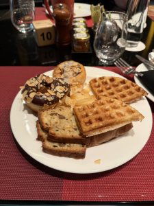 A white plate filled with a variety of breakfast pastries including a chocolate donut topped with sliced almonds, a light flaky pastry, and several pieces of waffle.