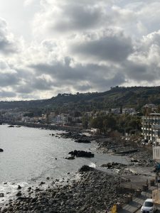 A sweeping view of the rocky Sicily coastline along the Mediterranean Sea, with coastal buildings nestled against green hills under a dramatic, cloudy sky.

