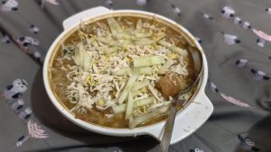 A white bowl filled with a savory snack called Chotputi, topped with shredded eggs, sliced cucumbers, and crispy crackers on a gray bird-patterned surface.
