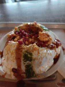 A close-up of a loaded Raj Kachori, topped with thick yogurt, green and tamarind chutneys, crunchy sev, and vibrant pomegranate seeds.
