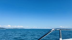 Bright landscape of a deep blue ocean under a clear sky, with distant green islands on the horizon, a small boat leaving a wake, and a chrome railing in the foreground.