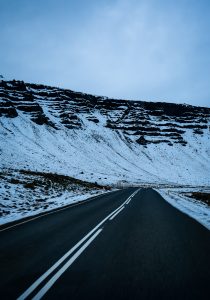 A winding road stretches through a snowy landscape, with steep, layered cliffs rising on one side. The sky is overcast, giving the scene a muted, cool tone. Snow covers the ground, blending with patches of grass visible along the roadside. Road markings are clear, guiding the way through this remote, natural setting.