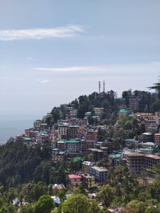 Dense hillside town with colorful buildings stacked along a slope, surrounded by trees. The scene overlooks a valley under a clear sky.