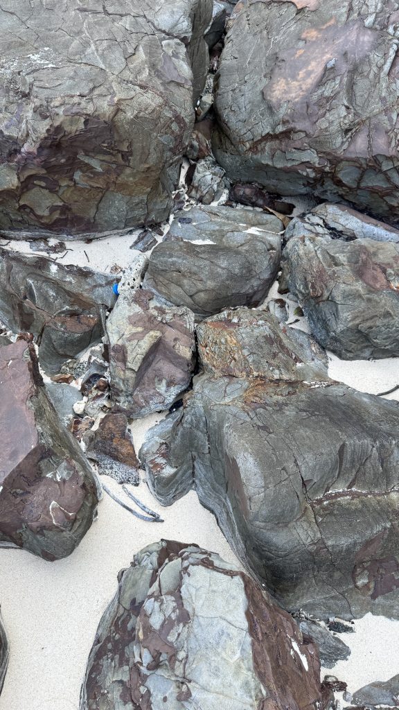 A top view shows several large and small rocks scattered on white beach sand. The rocks have a mix of grey and reddish brown colors with many natural cracks and lines on their surfaces. The sand is clean and visible between the different rock shapes.
