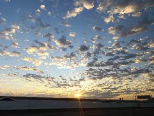 A vibrant sunset is visible over a highway, with a sky filled with scattered clouds in various shades of blue and gray. The sun is setting on the horizon, casting warm golden light that contrasts with the cool tones of the sky.