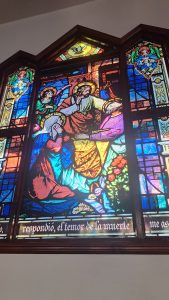 A colorful stained glass window depicting a religious scene. The central figure appears to be Jesus, engaged in a gesture with two other individuals, possibly conveying a message of compassion or healing. Surrounding them are angels and figures in vibrant robes. The window includes floral decorations and features inscriptions in Spanish along the bottom, providing context to the scene. The light filters through the glass, enhancing the vivid colors and intricate details.