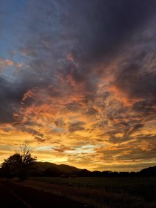
A vibrant sunset over a landscape featuring rolling hills. The sky is filled with dramatic clouds in shades of orange, yellow, pink, and purple, creating a breathtaking view.