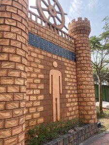 A brick tower wall with a stylized human figure and a large wheel on top. Small shrubs line the base under a partly cloudy sky, evoking a historical feel.