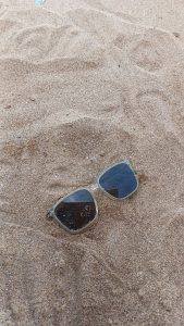 A pair of sunglasses rests in light brown sand, partially buried, with some sand particles visible on the lenses.
