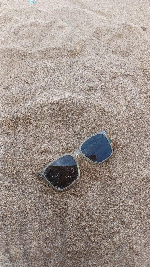 A pair of sunglasses rests in light brown sand, partially buried, with some sand particles visible on the lenses.