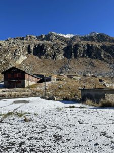 A snow-dusted landscape featuring a wooden cabin with red shutters in the foreground, a gravel path leading between the cabin and a small stone structure, and rocky mountains towering in the background under a clear blue sky. Switzerland