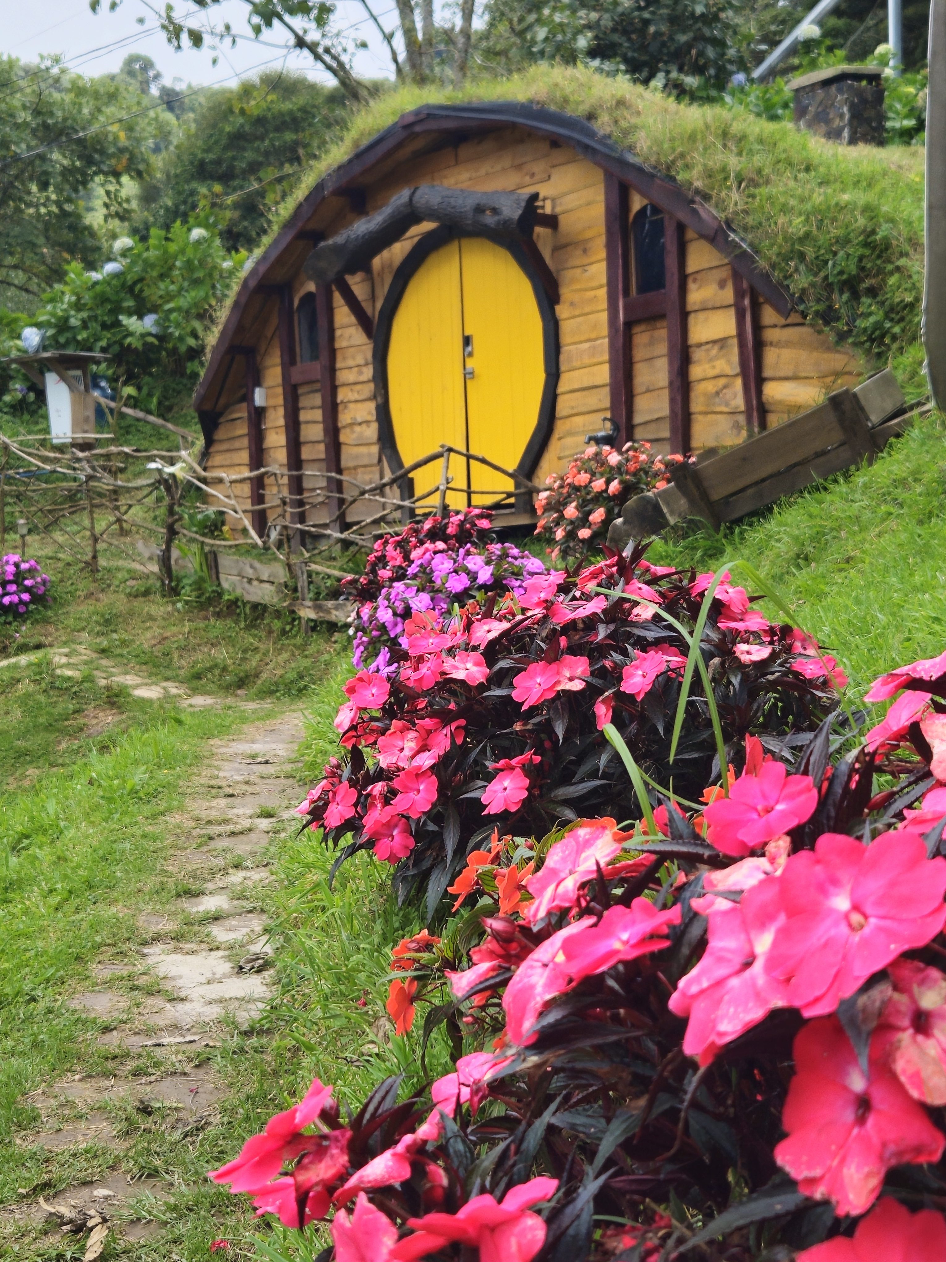 A charming hobbit house with a yellow door is nestled amidst vibrant pink and purple flowers, surrounded by lush greenery.