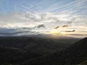 Sunset over rolling green hills and distant mountains, with scattered clouds in a partly cloudy sky, casting warm light across the landscape.
