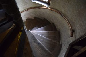 High-angle view looking down a narrow wooden spiral staircase with a curved handrail in a historic stone building.
