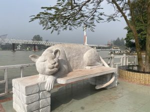 A large cat sculpture lies on a bench, appearing relaxed, near a riverside with a bridge in the background. Trees frame the scene under a clear sky.