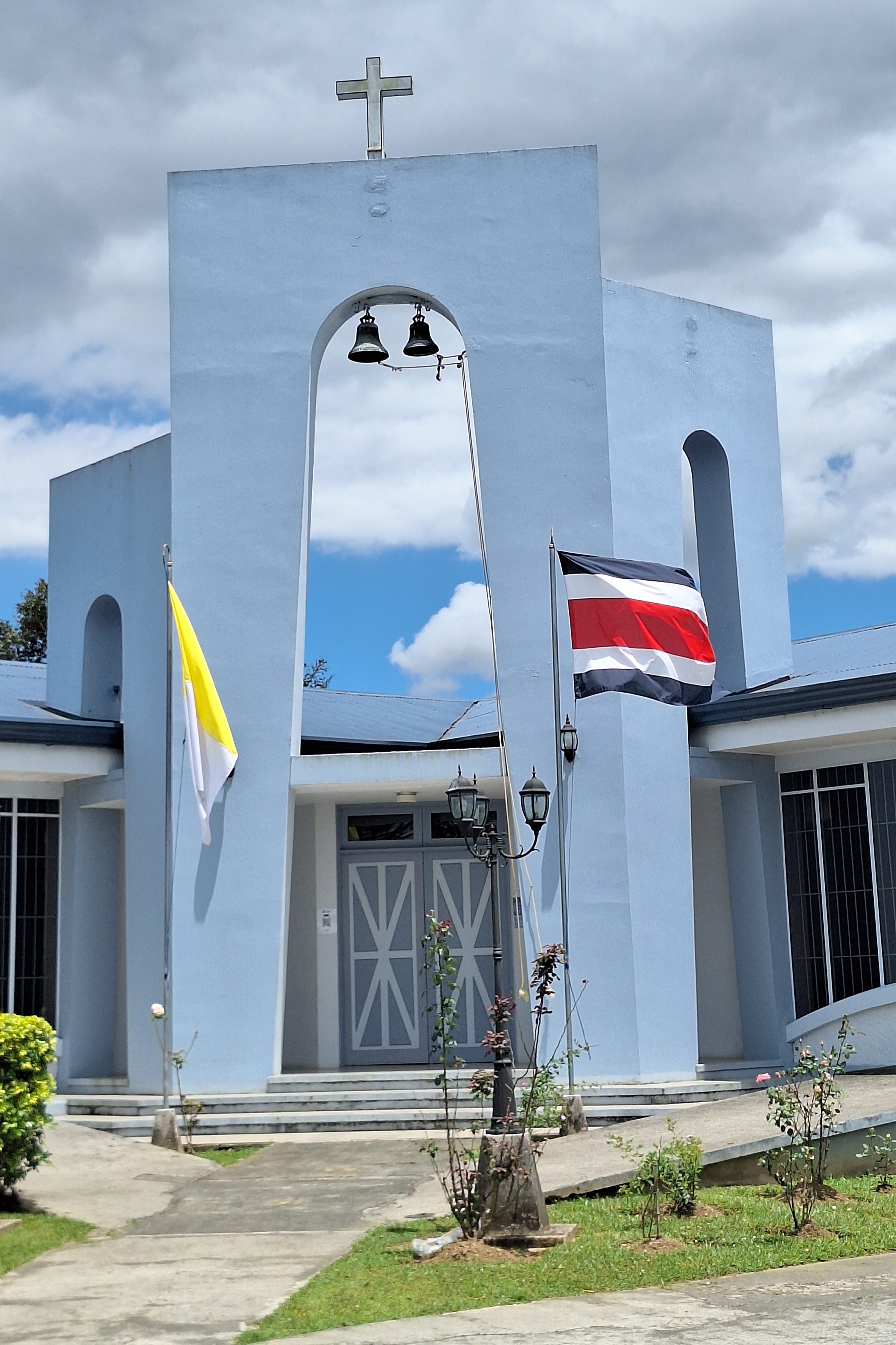 A sky-blue church with two black bells and a cross on top, with Vatican and Costa Rica flags in front, surrounded by pathways, greenery, and lampposts under a cloudy sky.