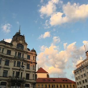 Ornate European buildings in Prague under a blue sky with sunset clouds. A Neo-Baroque facade stands beside modern buildings, with red-tiled roofs in the background. Warm evening light shines on the clouds and stone walls.