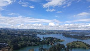 A panoramic view of rolling green hills and a serene lake, dotted with small islands and houses. The sky is bright blue with scattered white clouds. Trees line the banks of the lake, and some boats can be seen on the water. In the distance, more hills rise under a partly cloudy sky, creating a picturesque natural landscape.
