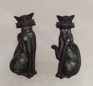 Two decorative cat figurines made from a dark material are displayed against a light background. The cat on the left has a green floral design along its body and appears to be sitting with a calm expression. The cat on the right features a bowtie and a similar floral pattern, sitting upright with one paw raised. Both cats have large, expressive eyes and detailed whiskers.