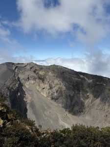 A panoramic view of a volcanic landscape featuring steep, rugged slopes of grey and brown rock. The summit shows signs of erosion, with loose debris scattered down the slopes. The sky above is partially cloudy, with patches of blue peeking through. In the foreground, some vegetation with green foliage is visible, contrasting against the stark terrain of the volcano.