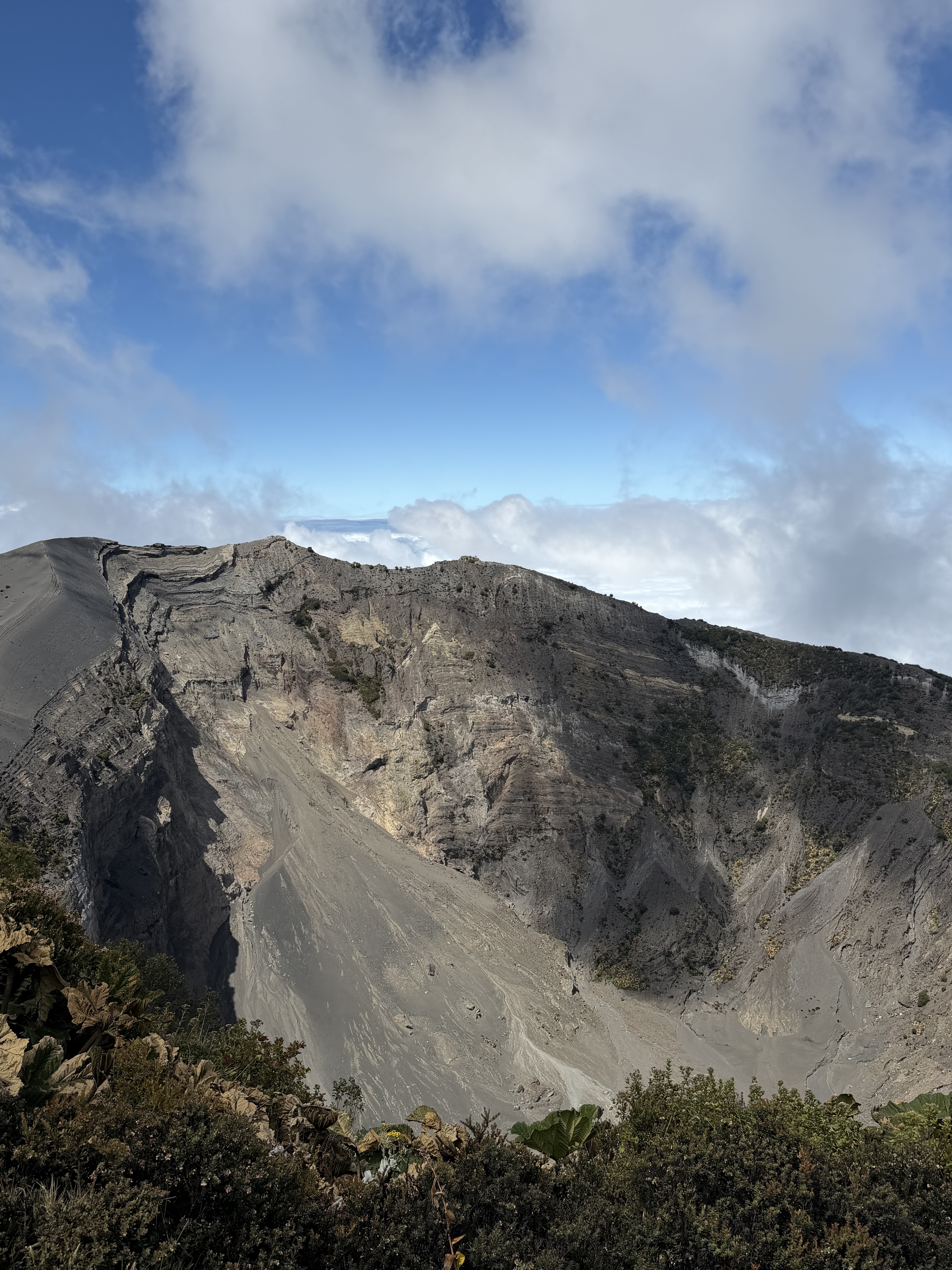 A panoramic view of a volcanic landscape featuring steep, rugged slopes of grey and brown rock. The summit shows signs of erosion, with loose debris scattered down the slopes. The sky above is partially cloudy, with patches of blue peeking through. In the foreground, some vegetation with green foliage is visible, contrasting against the stark terrain of the volcano.